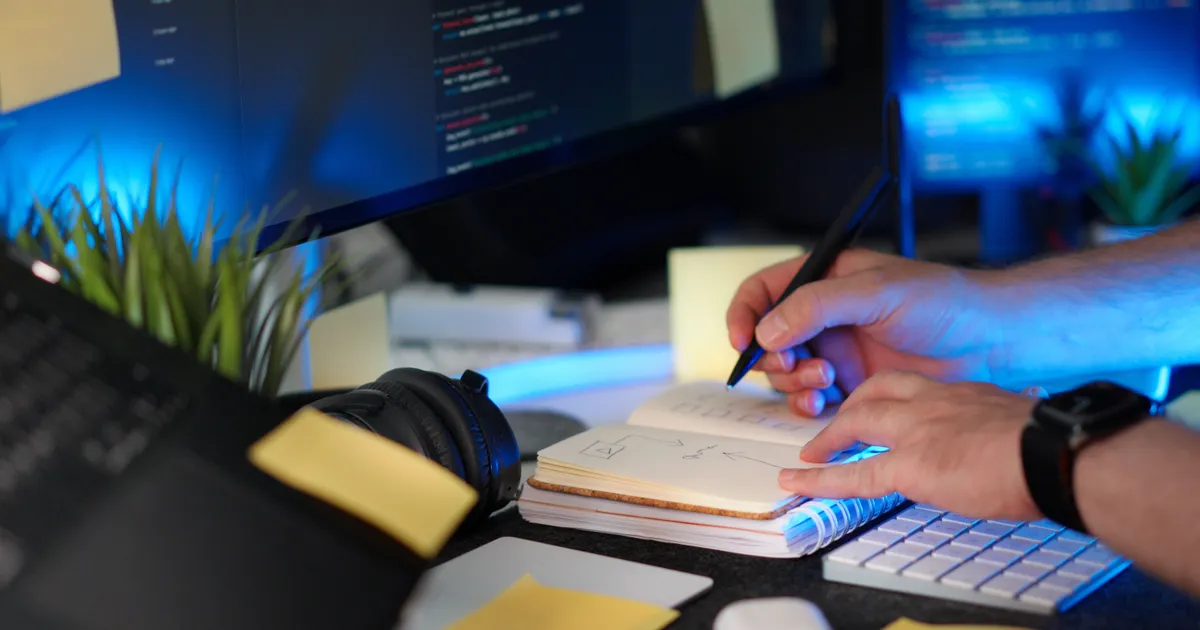 Desk with a code monitor, a notebook with a sketched diagram, and a hand writing notes in blue light